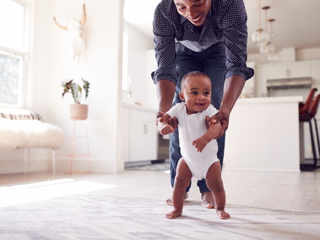 A toddler taking his baby first steps with his father’s help.